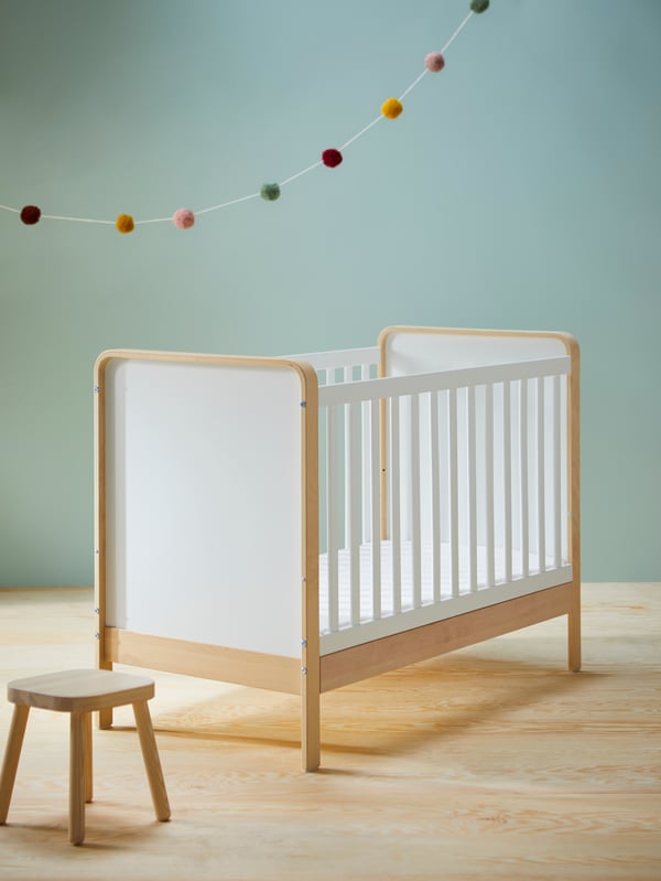 Un tabouret pour enfant FLISAT à côté d’un lit bébé ÄLSKVÄRD en bouleau et blanc, dans une pièce au sol en bois et aux murs gris/vert.