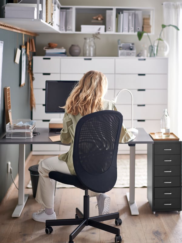 A woman sitting in a black FLINTAN office chair by a grey sit/stand RODULF desk with a computer, work lamp and drawer unit.