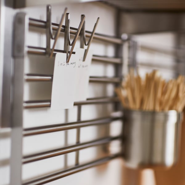 A white kitchen wall with a stainless steel KUNGSFORS wall grid with a KUNGSFORS container and pinned notes.