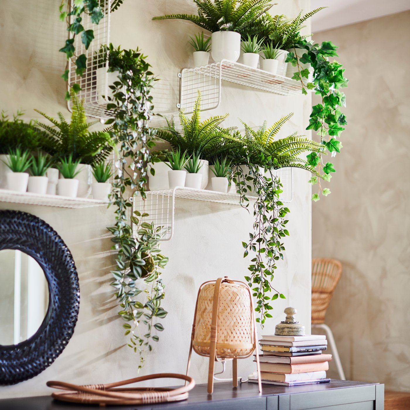 Sala con repisas ancladas a una pared de color blanco, con plantas sobre las repisas, abajo una mesa con una lámpara y libros sobre ella.
