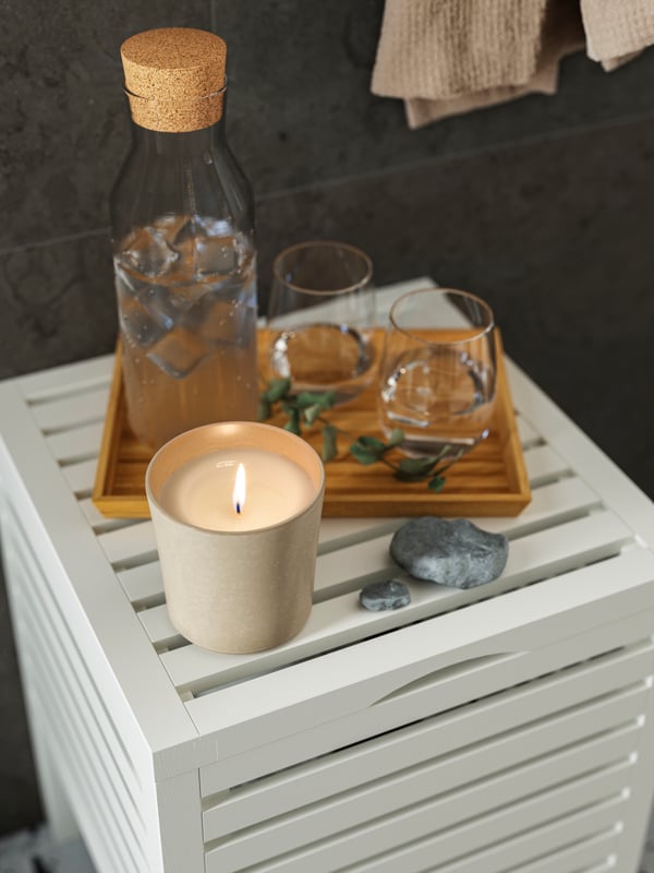 Le dessus d’un tabouret-rangement MUSKAN blanc avec un plateau sur lequel sont posés des verres d’eau, une bouteille et une bougie parfumée JÄMLIK allumée.