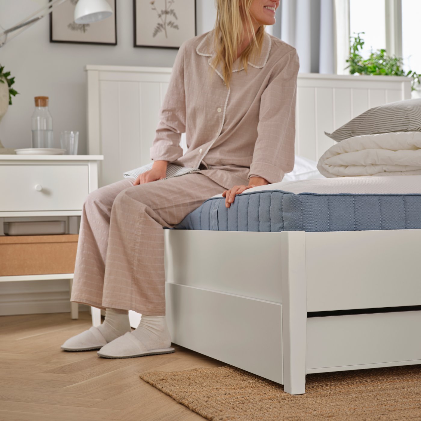 In a bedroom, a person in pyjamas, socks and slippers is sitting on a white GULLABERG bed frame with four storage boxes.