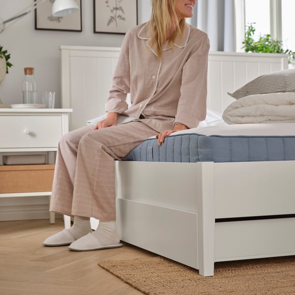 I ett sovrum sitter en person i pyjamas, strumpor och tofflor på en vit GULLABERG sängstomme med fyra förvaringslådor.