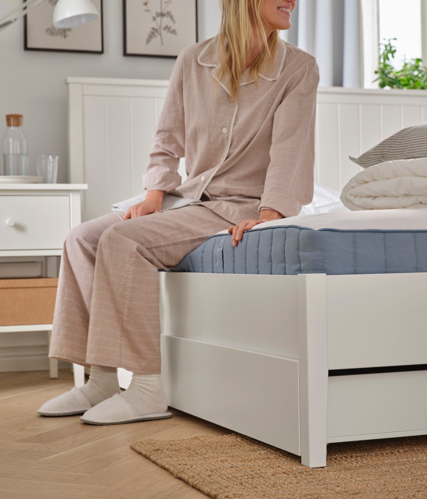 In a bedroom, a person in pyjamas, socks and slippers is sitting on a white GULLABERG bed frame with four storage boxes.