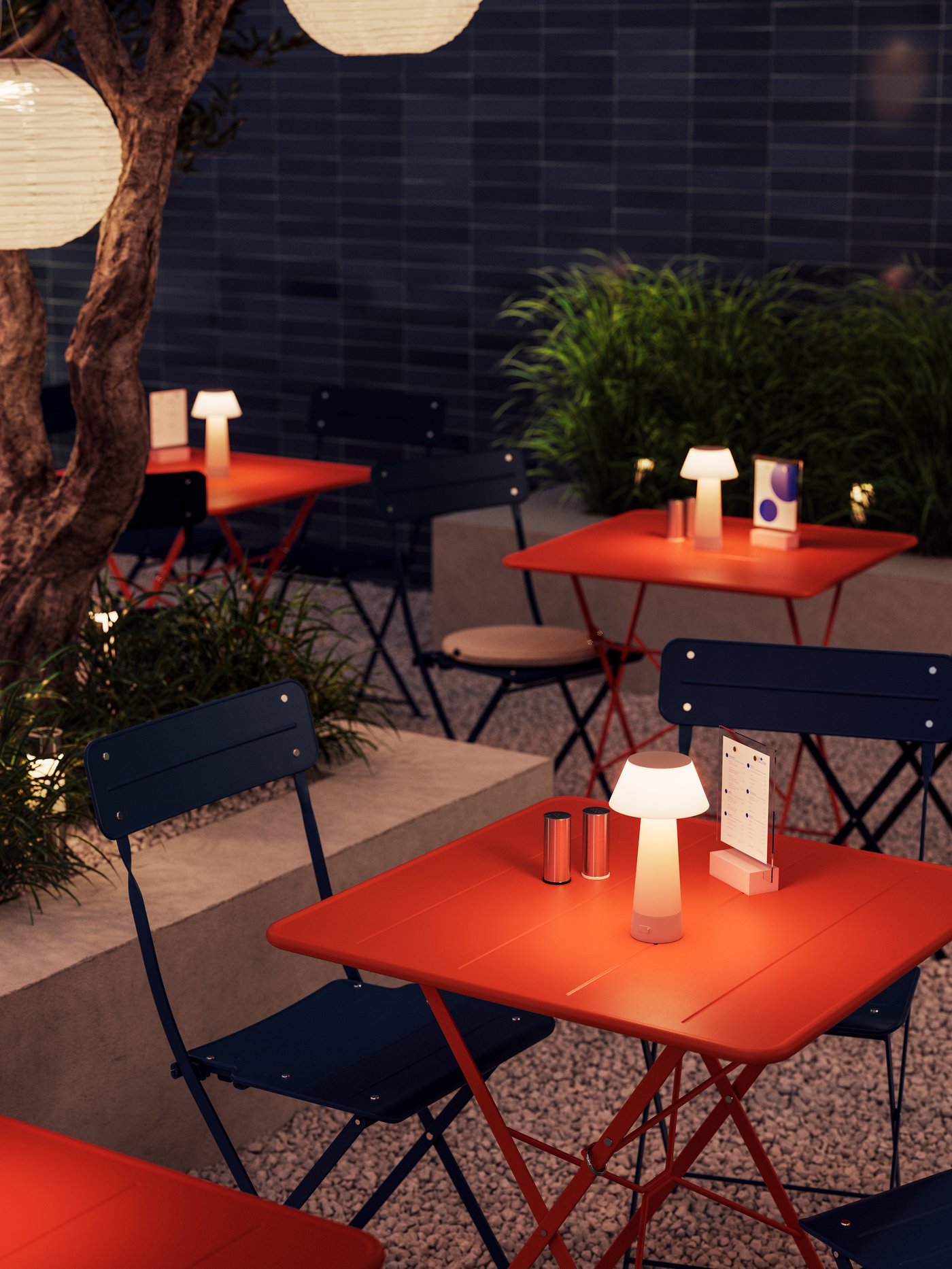 An outdoor cafè at sunset. Groups of SUNDSÖ outdoor bistro sets in dark blue and orange with cosy SOLVINDEN LED table lamps.