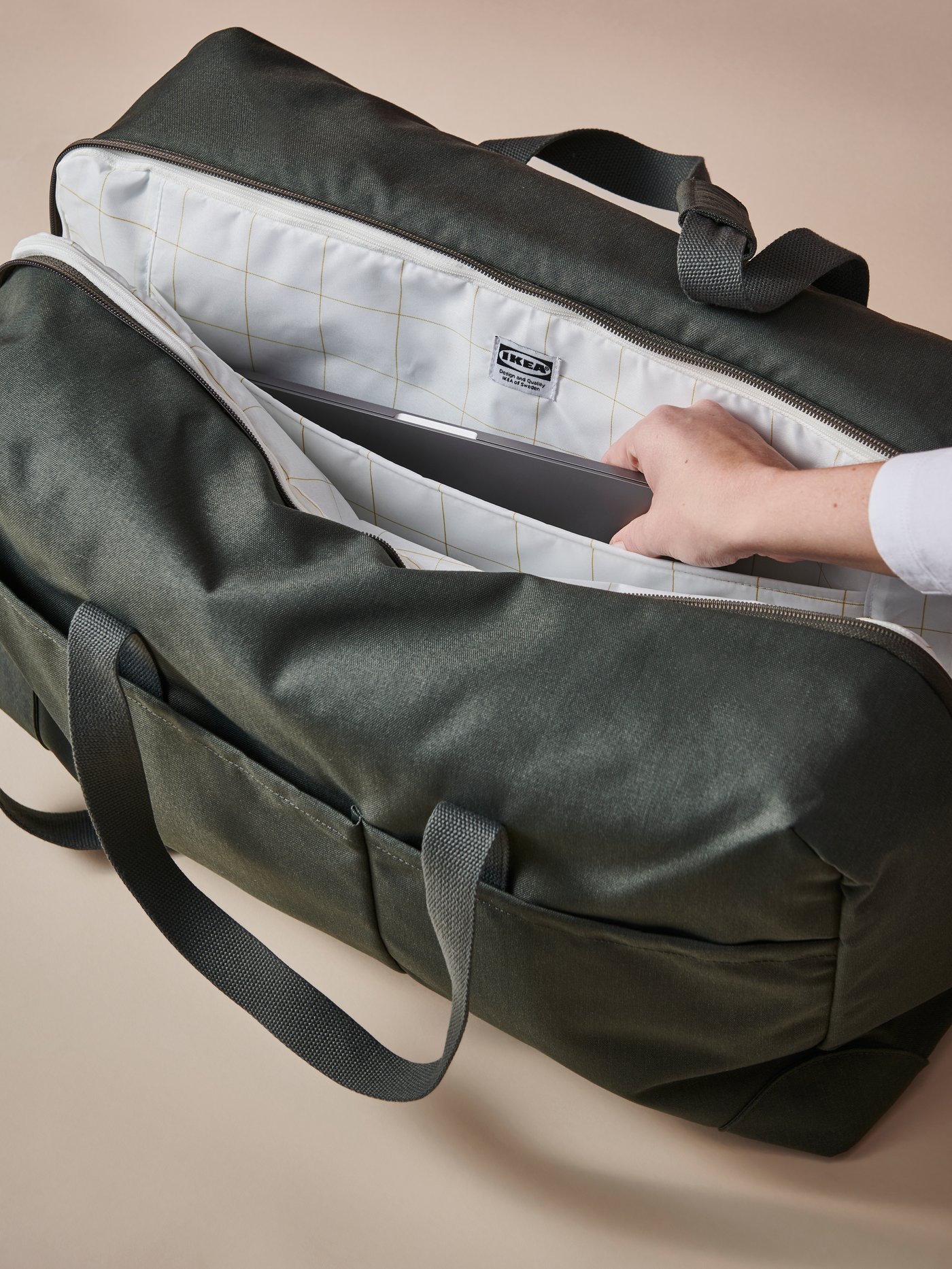 Une personne sortant un portable d’un sac de week-end DRÖMSÄCK vert. La doublure arbore un motif à carreaux et une étiquette IKEA.