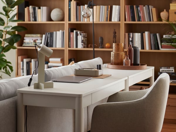 A living space with a small workstation placed behind a sofa, consisting of a white TONSTAD desk and a FLOTTILJ lamp.