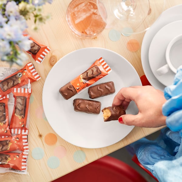 Une assiette blanche contenant des barres de chocolat au lait BELÖNING et une main tendue vers l'une d'elles.