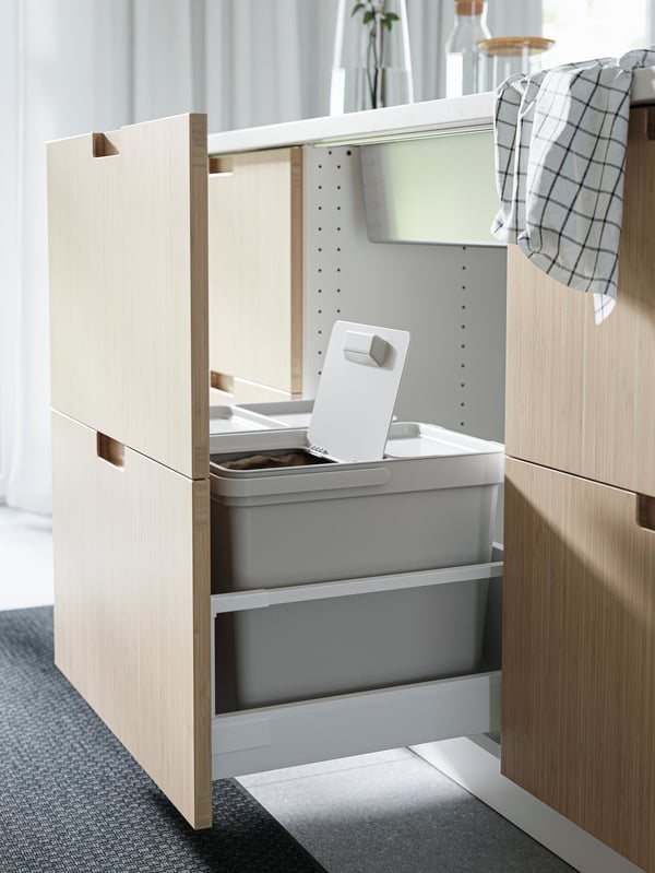 Kitchen drawer fronts in bamboo, light grey waste-sorting bins, a black rug, a tea towel in black/white and a white curtain.