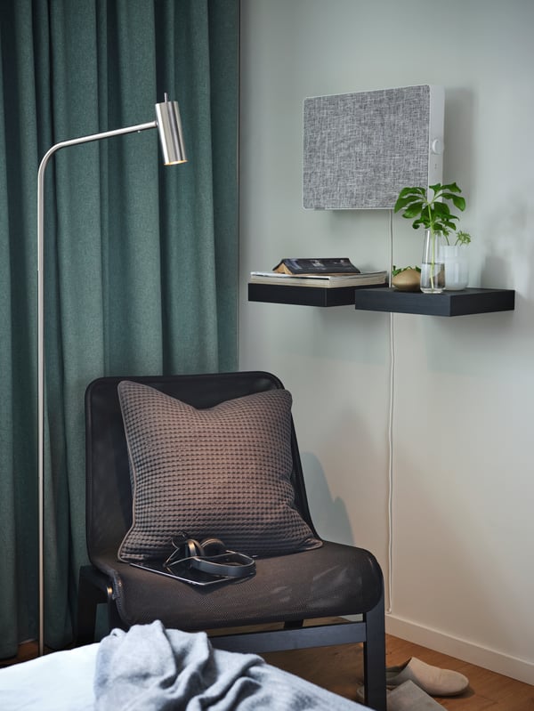 Una silla NOLMYRA con un cojin VÅRELD de apoyo. En la pared se ubica una repisa LACK con una planta y libros. A mano izquersa se posiciona una lampara de pie VIRRMO.