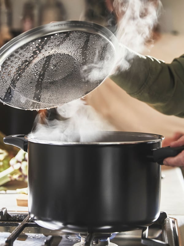 A person is holding the handle of a black HEMLAGAD pot with lid while opening the lid with the other hand to let out steam.