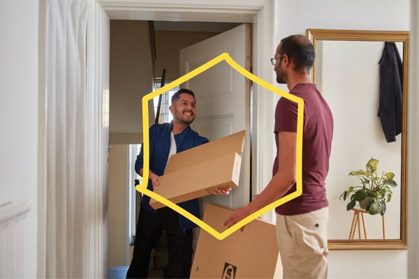 Dans un couloir lumineux, un homme reçoit des paquets plats IKEA de la part d'un livreur souriant.