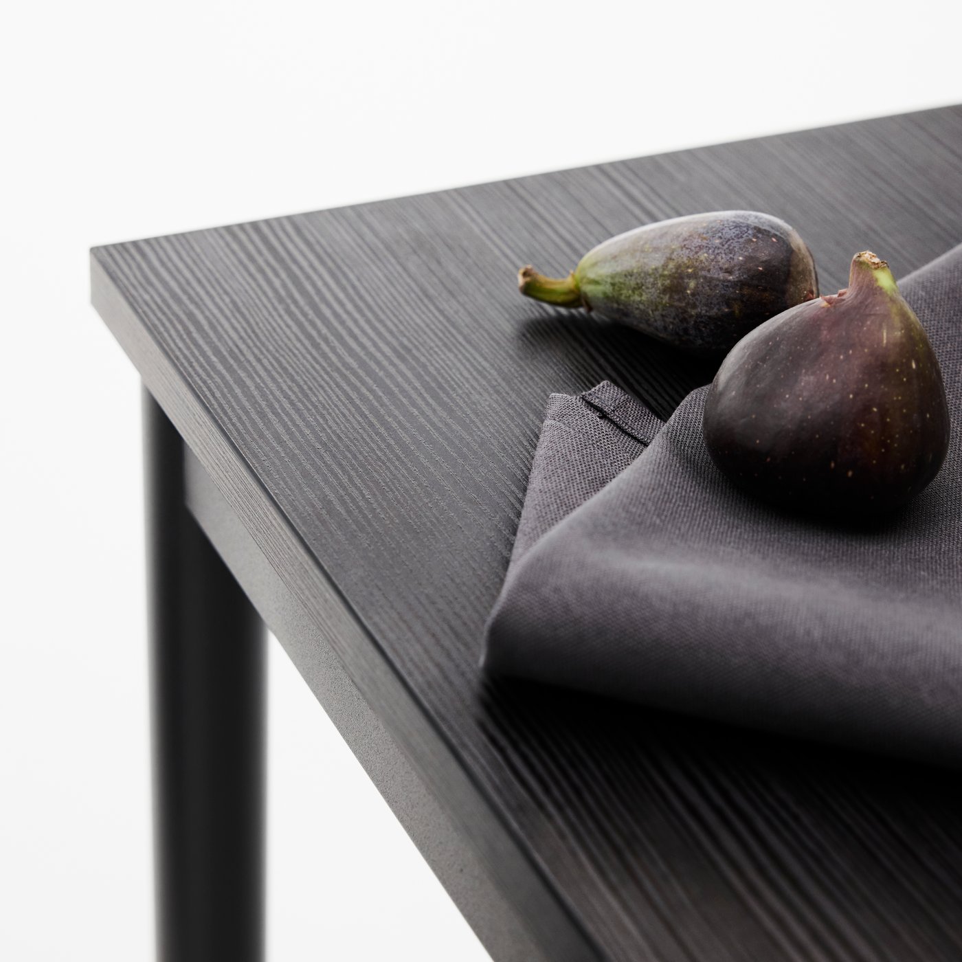 Gros plan sur le coin d’une table à manger SANDSBERG noire. Sur la table, une serviette noire et des figues.