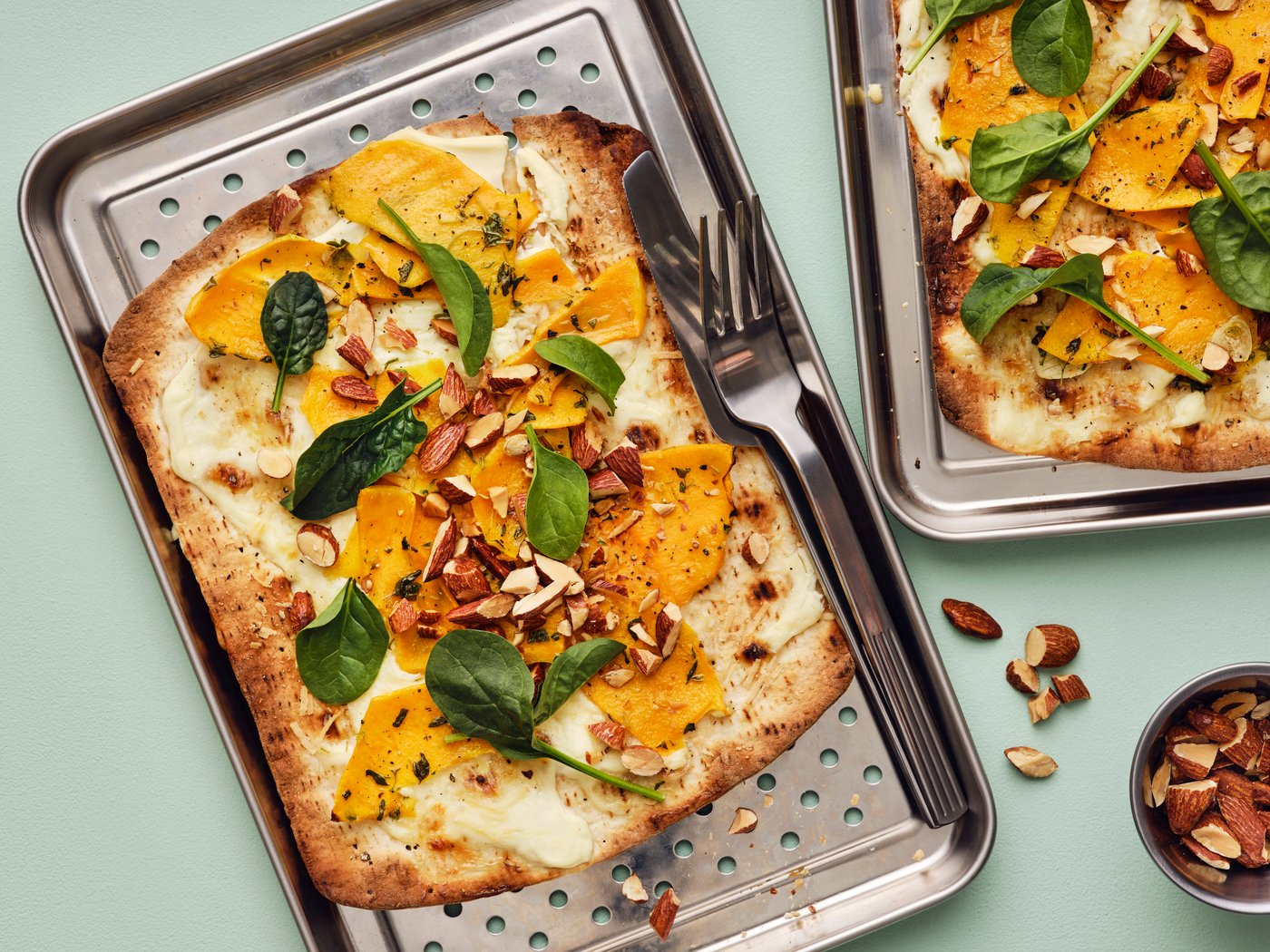 Twee pompoenpizza's met platbrood belegd met spinazie en MUNSBIT geroosterde amandelen op roestvrij stalen dienbladen met bestek erbij.
