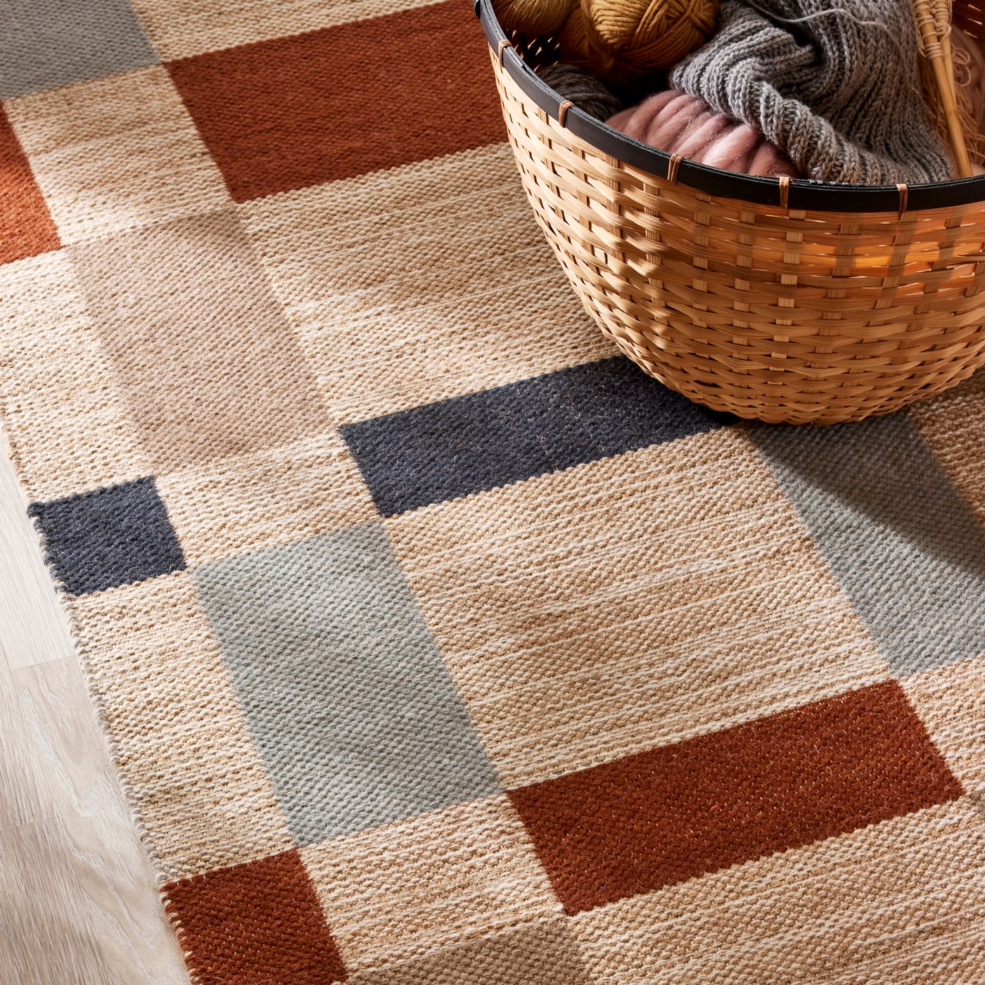 Un panier LÖVRÄFSA avec des tricots dedans est posé sur un tapis BLÅLJUS tissé à plat avec un motif à carreaux colorés.