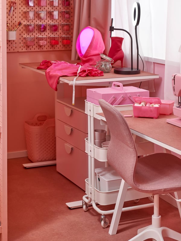 A desk with a chair is by the window to the right. On the pink wall in the back of the picture, there is a pink pegboard with pink accessories..