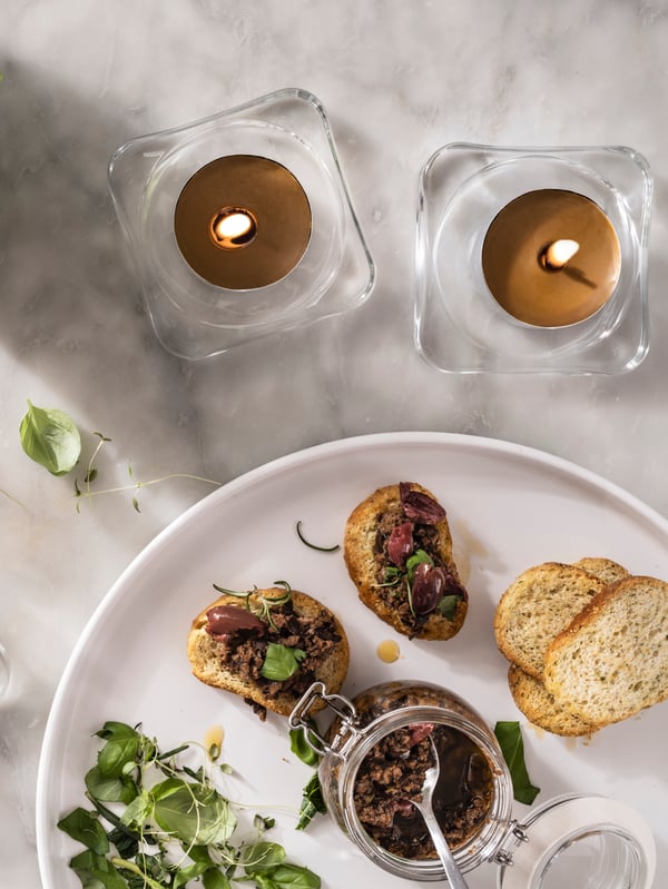 Two PRAKTRÖNN tea lights placed on GLASIG candle dishes amid canapés and tableware on a white-marble worktop.