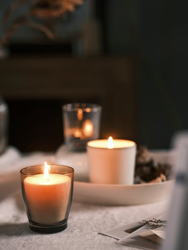A grey ENSTAKA scented candle, a white ADLAD scented candle on a white dish, plus a vase on a table with white tablecloth.