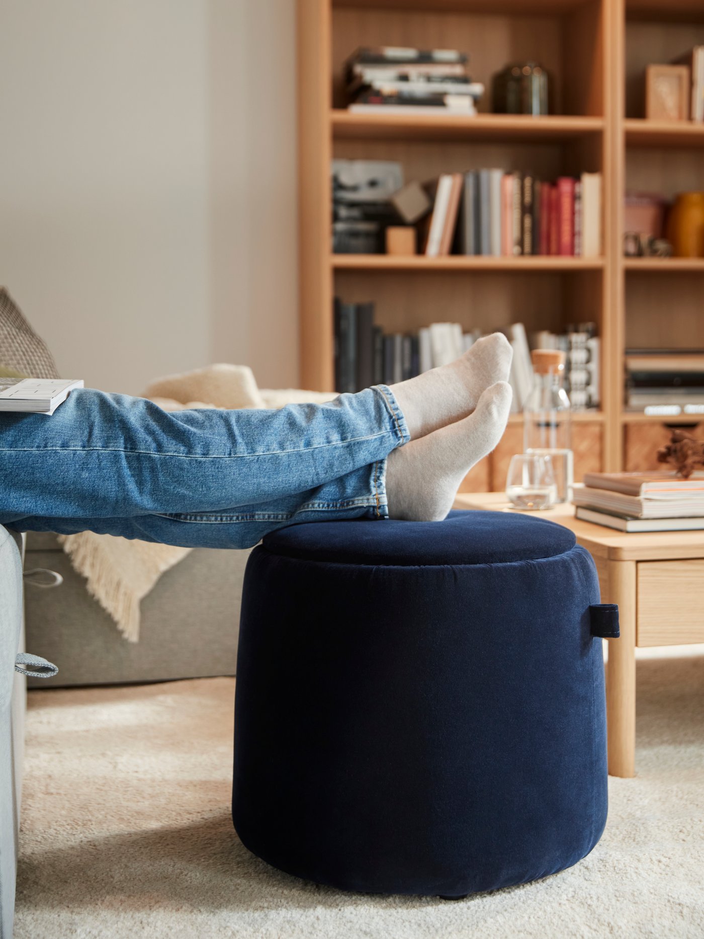 A person’s feet rest on a dark blue JÄRRESTAD pouffe with storage near a TONSTAD bookcase and a TONSTAD coffee table.