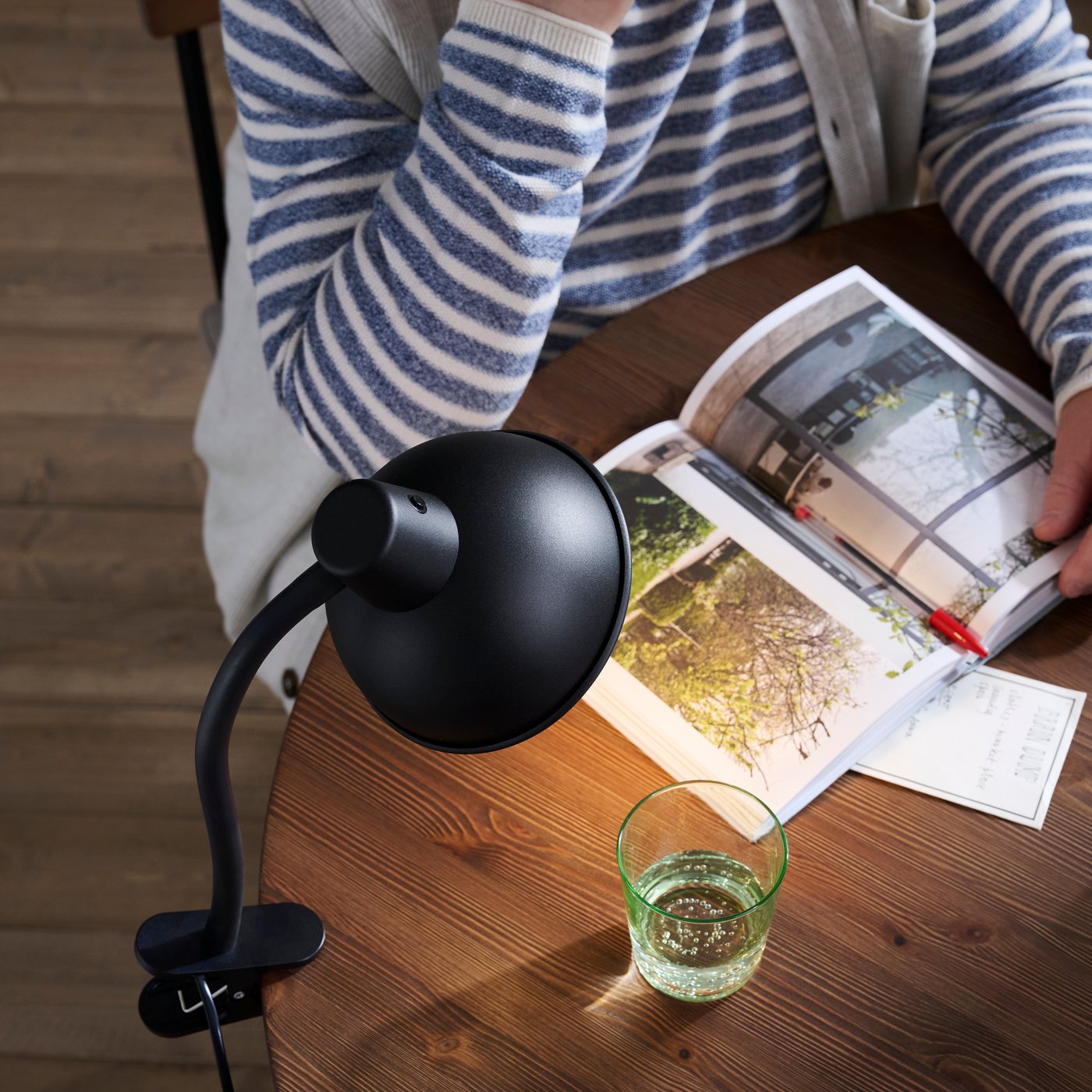A SKURUP clamp spotlight attached to a wooden table, shown beside a green glass, open magazine and person reading.