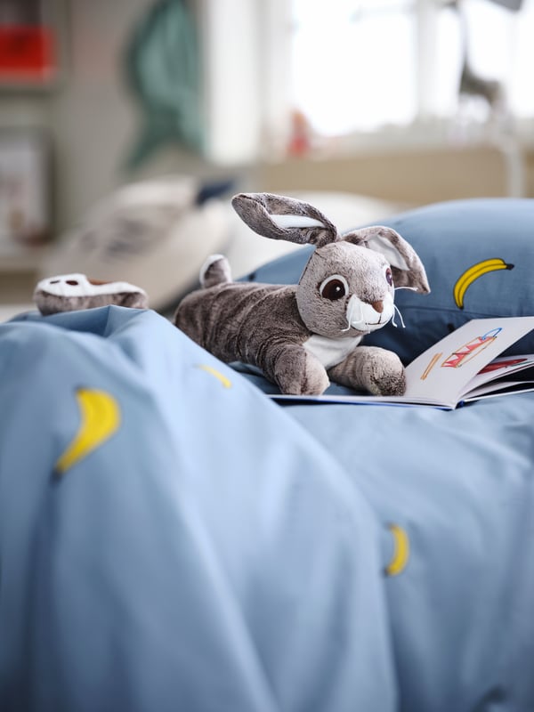 On a fluffy patch of a blue banana-pattern VÄNKRETS quilt cover lies a VANDRING HARE soft toy, seemingly engrossed in a book.