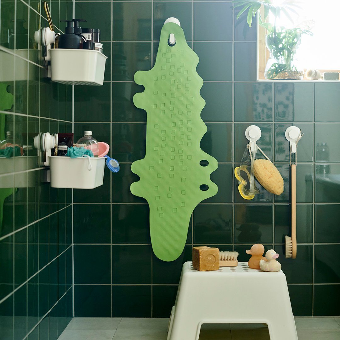 Un piso de baño en forma de cocodrilo PATRULL verde, colgado deun gancho TISKEN blanco adherido con una ventosa a un muro de baño con baldosas cuadradas de color verde botella. En el muro lateral se ven dos canastos TISKEN blancos y en el suelo un piso BOLMEN con artículos de baño. 