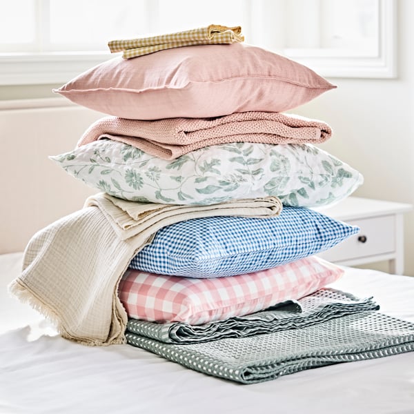 Pillows, throws and duvet covers, in different colours and patterns, stacked on top on a bed in a bedroom.