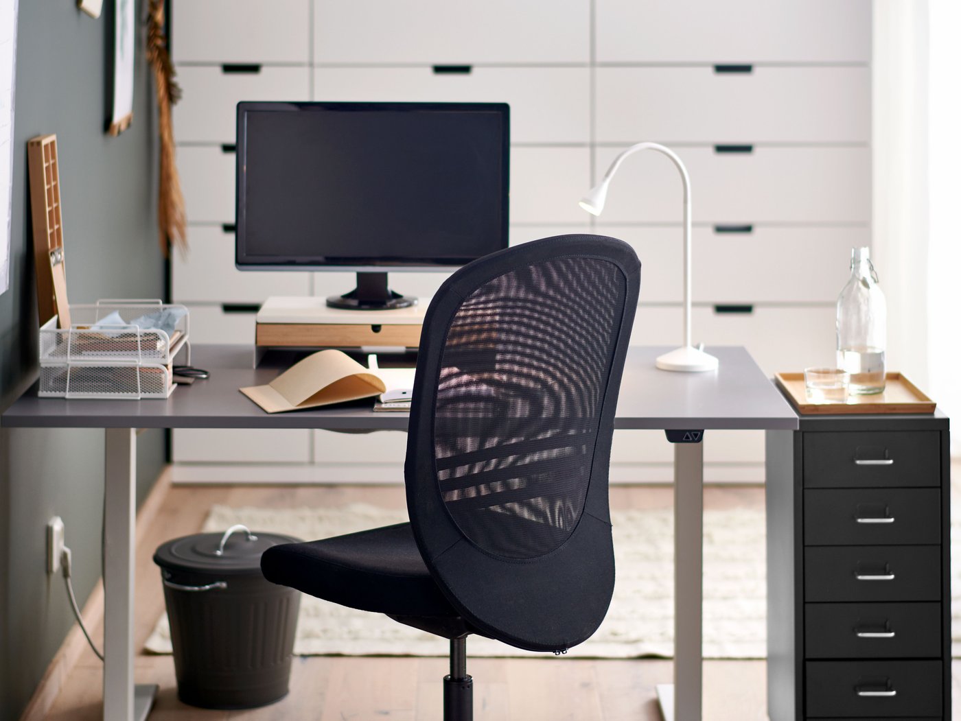 A work station with RODFUL sit/stand desk in grey/white and a black mesh office chair faces a wall of white cabnets.