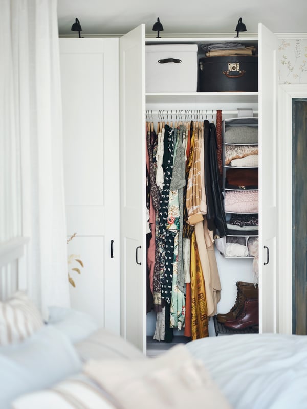 A PAX/GRIMO wardrobe in front of a double bed with two doors open, showing hanging and folded clothes and storage boxes.