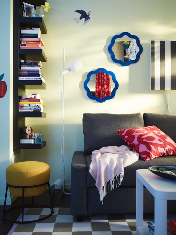 Un angolo colorato con una mensola a muro LACK nero-marrone con pile di libri, un divano-letto FRIHETEN e un pouf giallo EMMISLÖV.