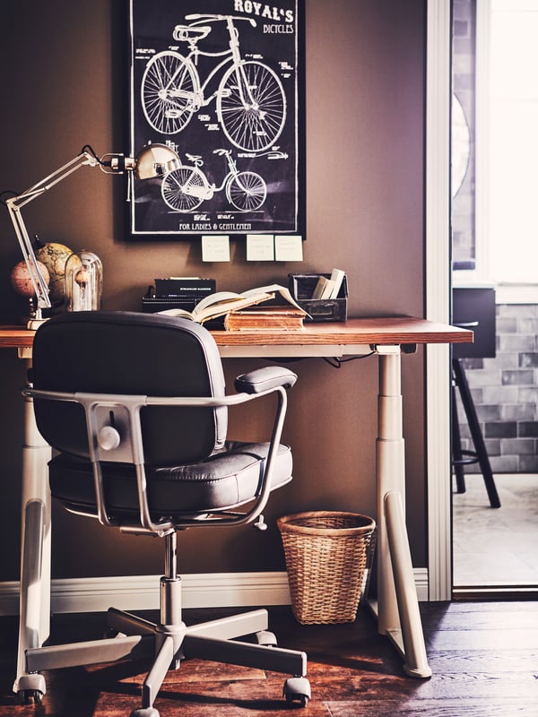 Un ambiente de homeoffice en el que se ve una silla ALEFJÄLL negra detrás de un escritorio IDÄSEN café, de fondo en la muralla hay un cuadro de bicicletas y en el piso un basurero TRUMMIS.