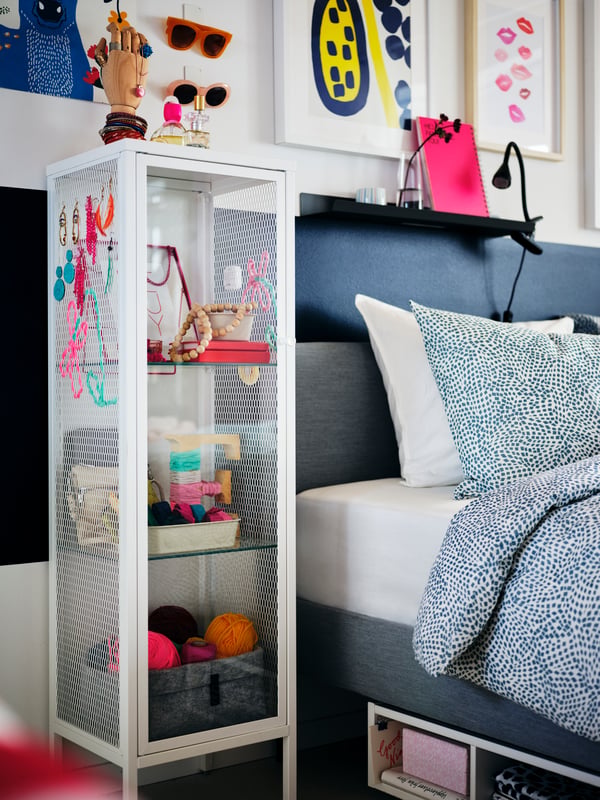 A white BAGGEBO glass-door cabinet holding various colorful items by a light-grey SLATTUM bed with TRÄDKRASSULA bed linen.