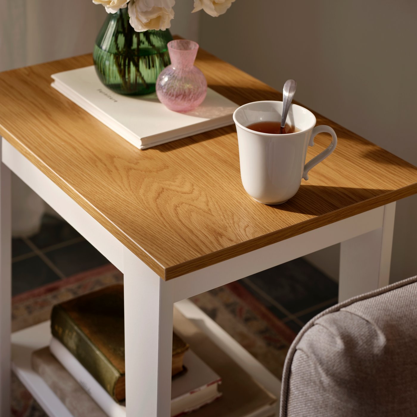A softly sunlit, white ÄMMARYD side table holding a white UPPLAGA mug with tea, a flower vase, and books on the lower shelf.