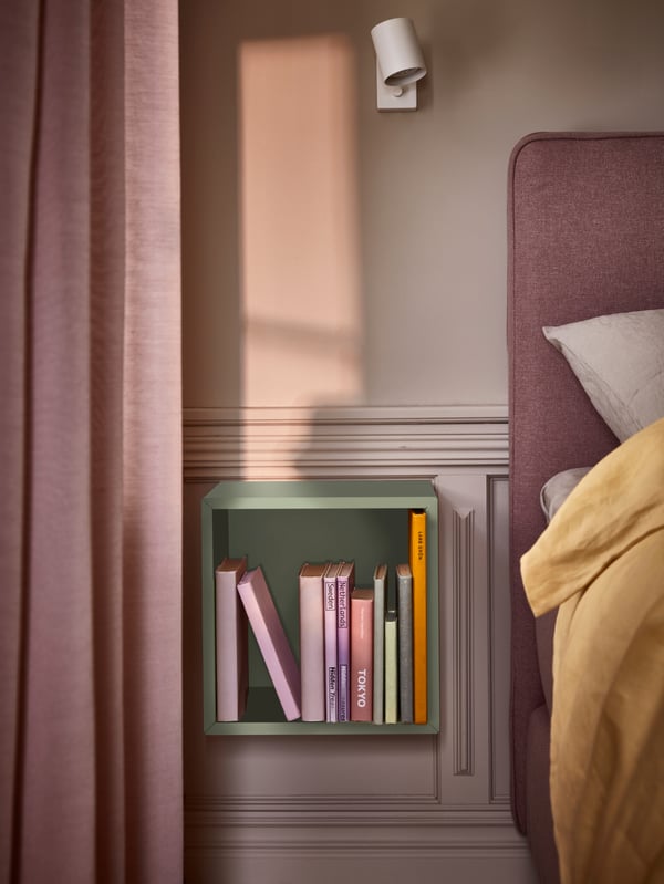 Une armoire EKET vert clair remplie de livres est fixée à un mur lambrissé, près duquel se trouvent un lit et des rideaux rosés.