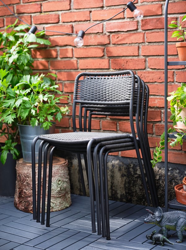 Four dark grey VIHOLMEN chairs stacked against a red brick wall with plants in terracotta pots on shelving to one side.