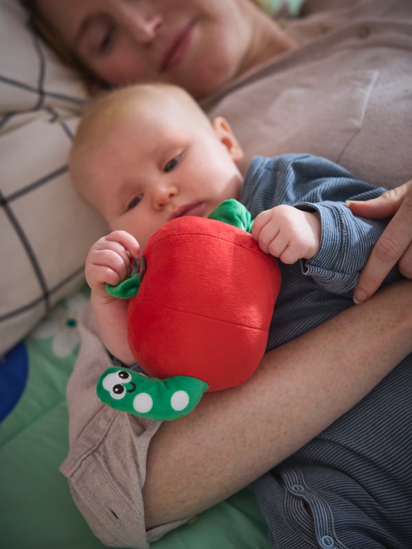 En baby, der ligger i sin mors arme og holder et rødt SANDBY æblelegetøj med musiklyde.