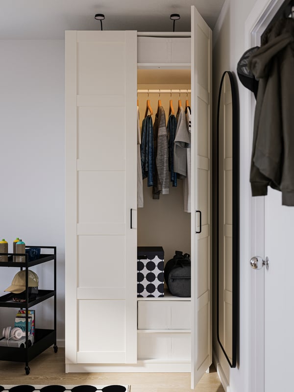 Armario PAX blanco con una puerta abierta que muestra la ropa ordenada en el interior junto a un carrito negro con ruedas a la izquierda.
