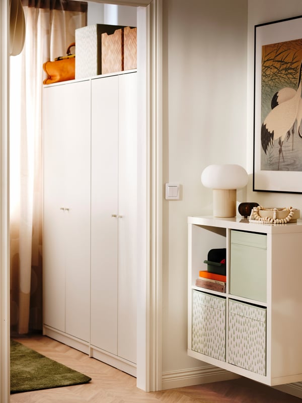 A high-gloss white KALLAX shelving unit is mounted to the wall in a room beside a bedroom with two KLEPPSTAD wardrobes.