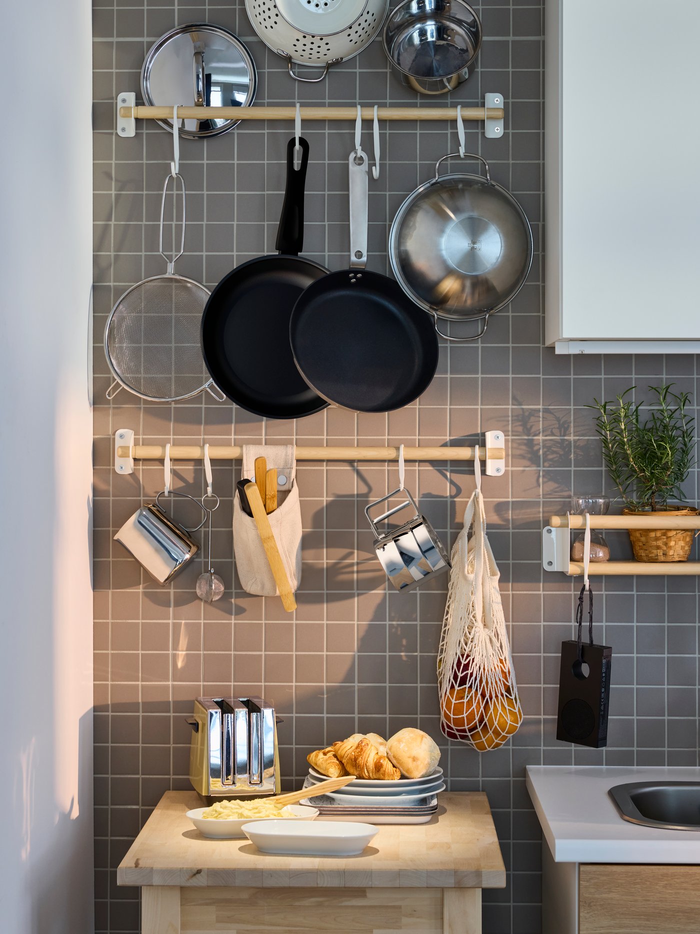 Una pared de cocina con dos barras KUNGSFORS con artículos de cocina como sartenes, coladores y jarros colgando de las barras.