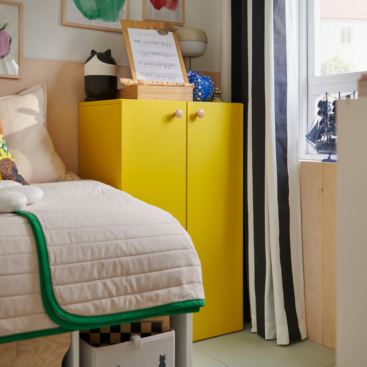 A bright yellow LASTARE storage combination with a child’s toys on top, sits beside a bed with a white and green bedspread.