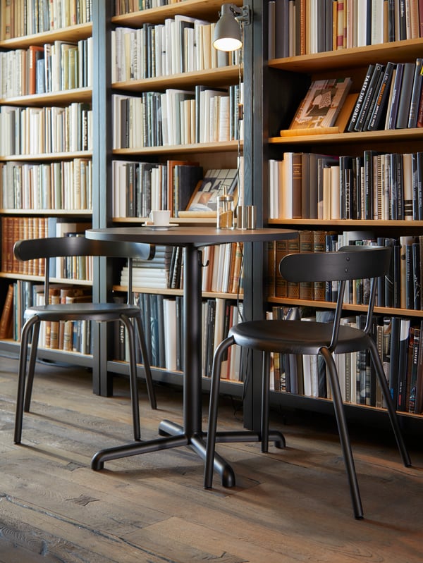 Round anthracite café table with hooks for bags underneath and two chairs, in front of a large bookcase with books.