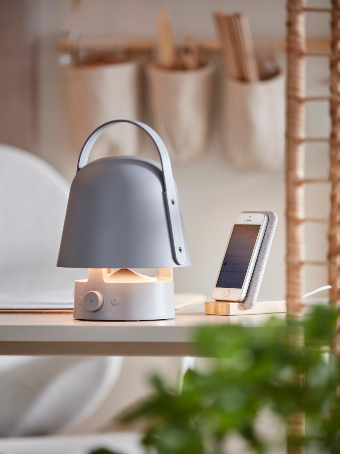 Lampada/cassa Bluetooth VAPPEBY grigia su una scrivania bianca, accanto a un supporto di ricarica wireless NORDMÄRKE in bambù.