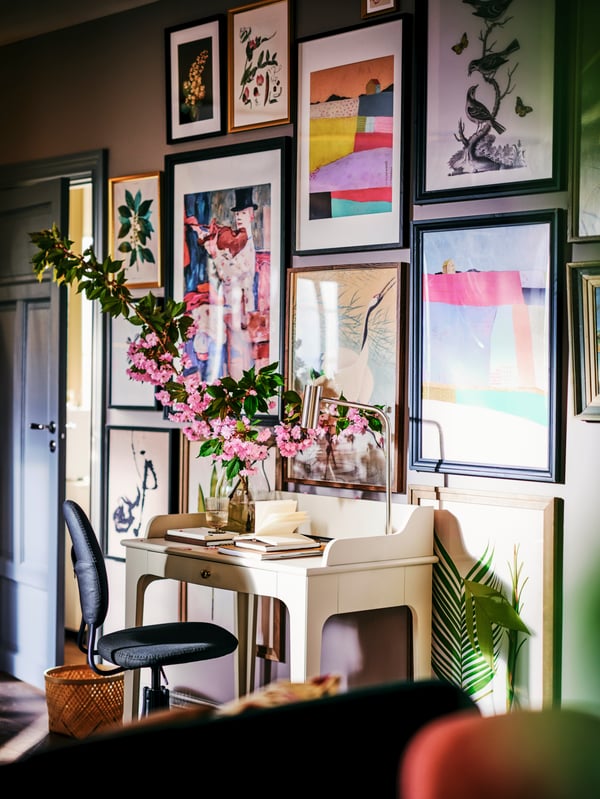 An IKEA LOMMARP off-white desk in a traditional style with flowers on top in a vase,  in front of a wall covered with a gallery of picture frames of various sizes and colours