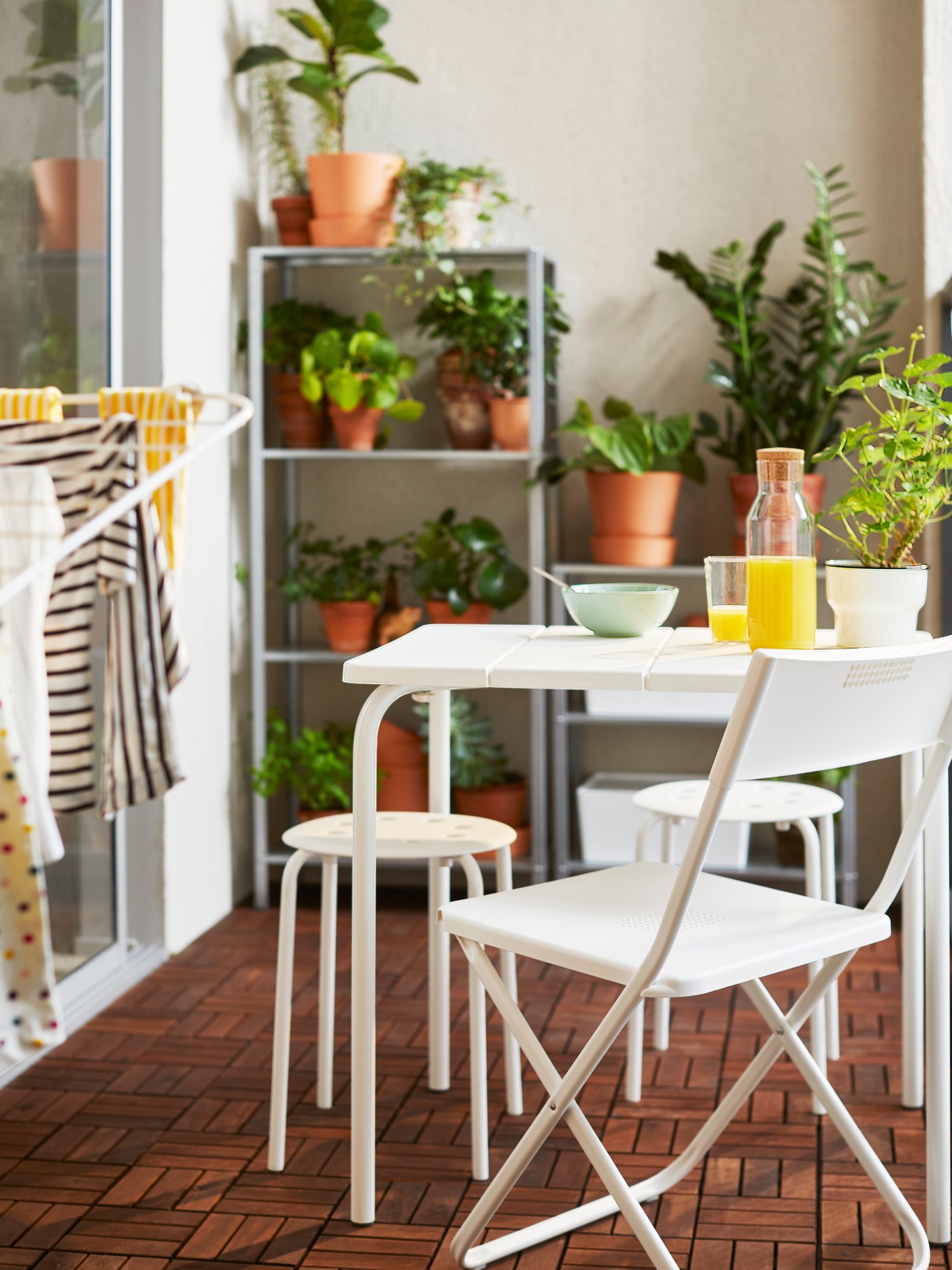 Una terraza con piso de madera donde se aprecia un juego de terraza con comestibles sobre la mesa, detrás un estante con plantas y al costado del comedor de terraza un tendedero de ropa.