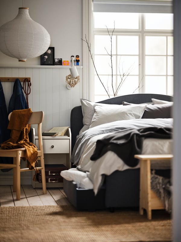 A room with a large bed by a window, grey BLÅVINDA bed linen, a white HAUGA bedside table and a white RISBYN pendant lamp.