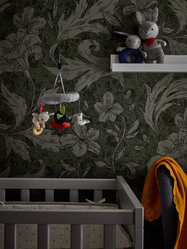 Un mobile GULLIGAST suspendu au-dessus d’un lit enfant placé près d’un mur à motifs floraux avec une bordure sur laquelle se trouvent un hochet et une peluche.