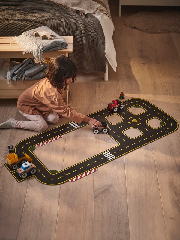 A child playing with a car on a RÄVUNGE car track set on a bedroom floor with a PERJOHAN bench with storage behind them.