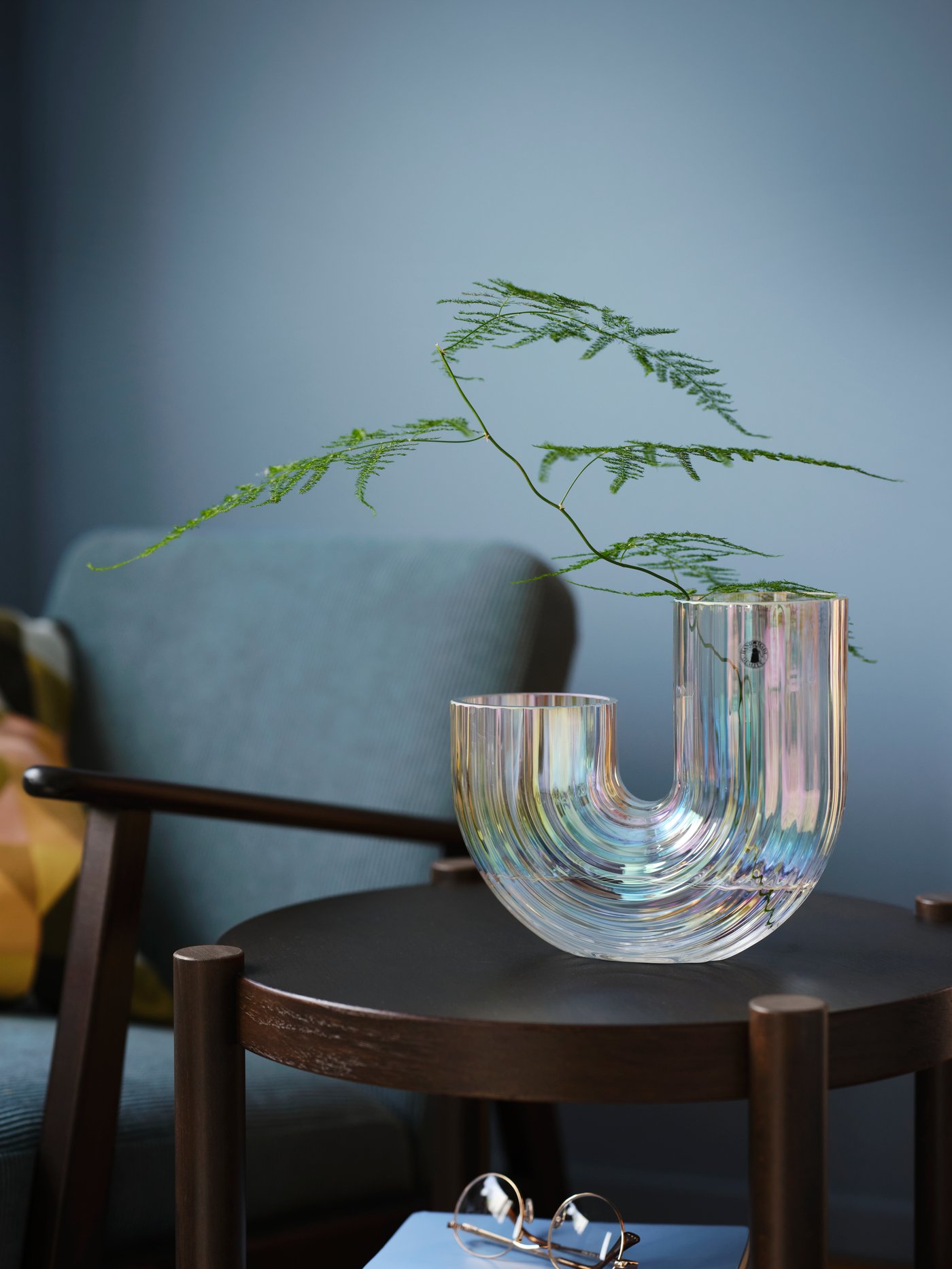 A mother-of-pearl-coloured RÄFFELBJÖRK glass vase holding a leafy twig stands on a dark-brown LISTERBY side table.