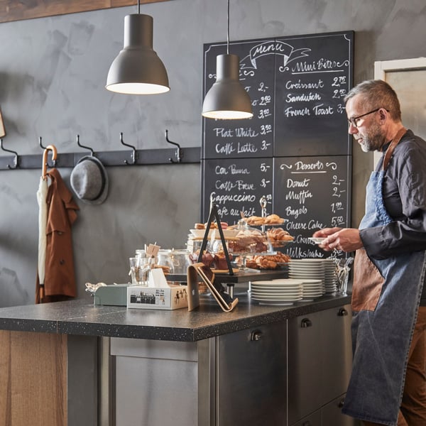 Person bakom cafédisk med bakverk och menytavla i en modern kafémiljö.
