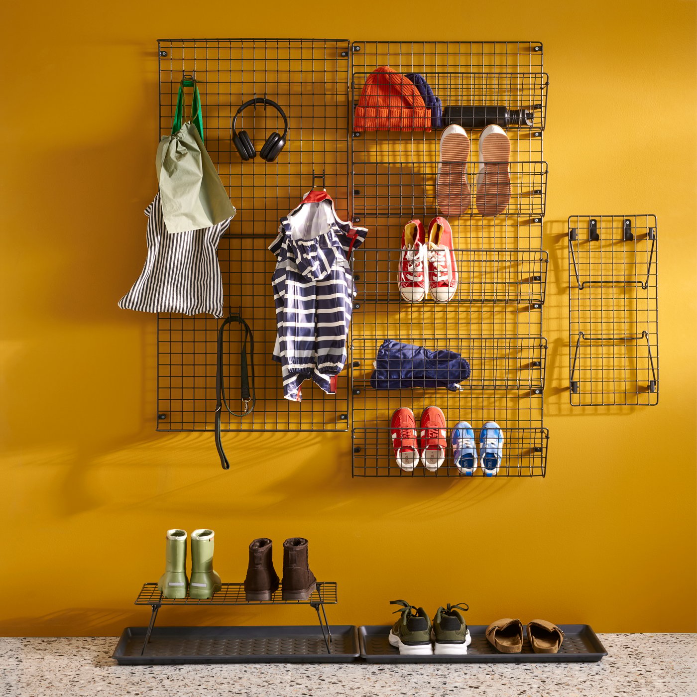 Black mesh GREJIG wall pockets filled with shoes and coats against a yellow wall with shoes on mats below.